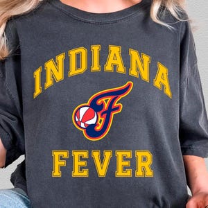May include: Dark gray t-shirt with the text "INDIANA FEVER" in yellow block letters. The shirt features a stylized logo with a basketball and the letter "F". The shirt is a comfortable, casual style.