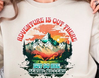 May include: A beige sweatshirt with a graphic print of a mountain range and the text "Adventure is out there, but so are serial killers".