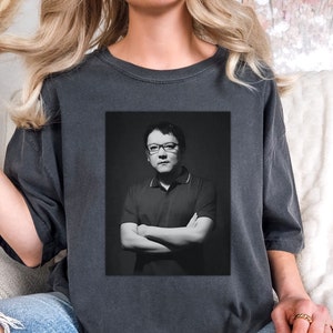 May include: A dark gray t-shirt with a black and white photo of a man wearing glasses and a collared shirt. The man is standing with his arms crossed.