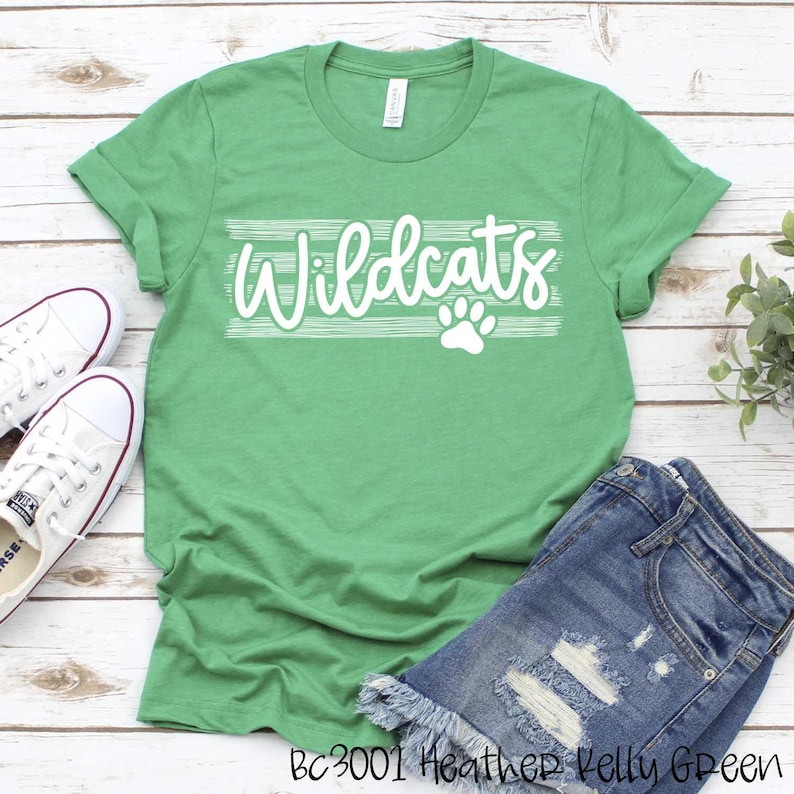 Op de afbeelding: Groen T-shirt met witte tekst die "Wildcats" zegt met een pootprint eronder. Het shirt ligt op een houten oppervlak met een paar witte sneakers en een blauwe spijkerbroek.