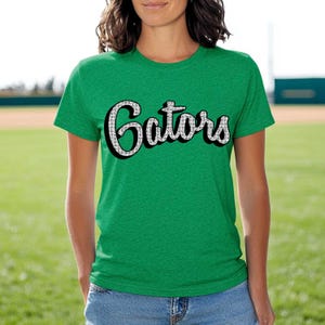 May include: Green t-shirt with the word "Gators" in a white, retro font with a black outline. The shirt is a crew neck style and is worn by a person. The background shows a baseball field.