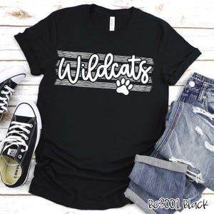 Op de afbeelding: Zwart T-shirt met witte tekst die "Wildcats" zegt met een pootprint eronder. Het shirt ligt op een houten oppervlak met een paar zwart-witte sneakers en een blauwe jeans.