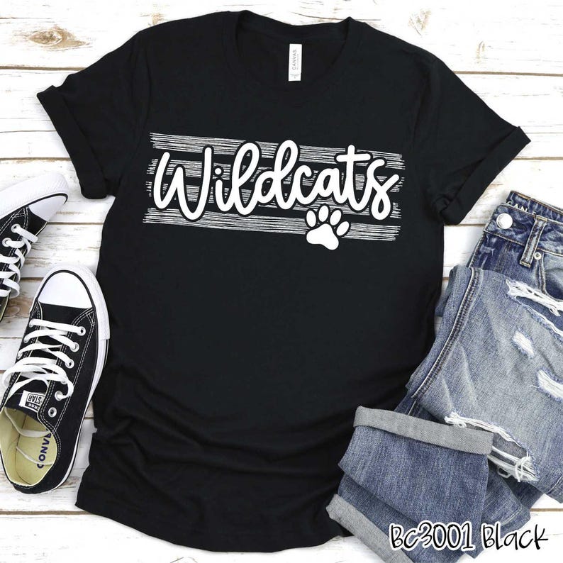 Op de afbeelding: Zwart T-shirt met witte tekst die "Wildcats" zegt met een pootprint eronder. Het shirt ligt op een houten oppervlak met een paar zwart-witte sneakers en een blauwe jeans.