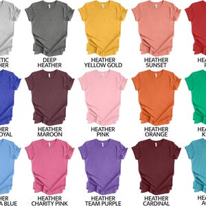 Op de afbeelding: Een kaart met 15 verschillende gekleurde T-shirts met gekrulde randen. De kleuren omvatten atletisch grijs, donker grijs, heide geel goud, heide zonsondergang, heide rood, heide koninklijk blauw, heide mahonie, heide roze, heide oranje, heide kelly, heide carolina blauw, heide liefdadigheid roze, heide team paars, heide kardinaal en heide aqua.