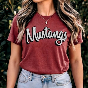 May include: A maroon t-shirt with the word "Mustangs" in a white, outlined, 3D font. The shirt is short-sleeved and made of a soft material. The model is wearing blue jeans and a gold necklace.