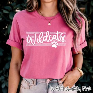 Op de afbeelding: Roze T-shirt met witte tekst die "Wildcats" zegt met een pootprint eronder. Het shirt heeft een ronde hals en korte mouwen.
