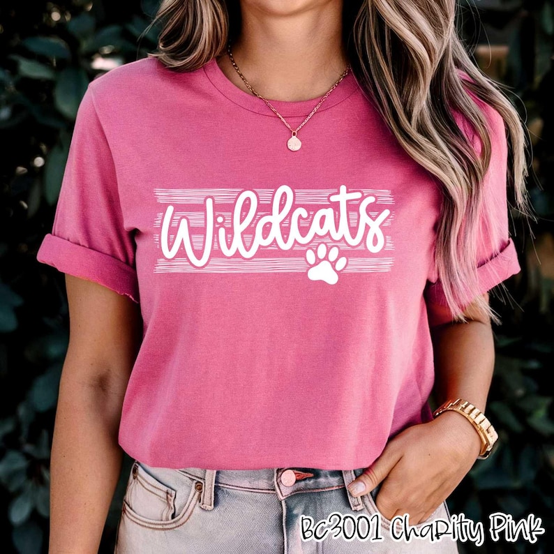 Op de afbeelding: Roze T-shirt met witte tekst die "Wildcats" zegt met een pootprint eronder. Het shirt heeft een ronde hals en korte mouwen.