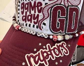 May include: A maroon and white trucker hat featuring a white front panel with a maroon bow, a football graphic, and the text "game day" and "GD". The brim is maroon with the word "raptors" in white and maroon, accented by beads spelling "CHEER MOM".