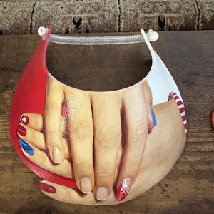 May include: A white visor with a red, white, and blue design featuring two hands with painted nails. The hands are holding each other.