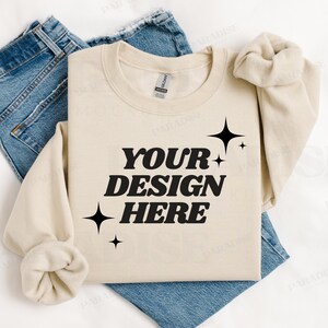 May include: A beige crewneck sweatshirt with the text "YOUR DESIGN HERE" in bold black letters, accented by starburst graphics. The sweatshirt is laid on top of blue denim jeans, creating a casual, everyday look.