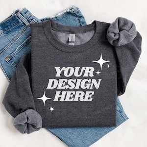 May include: A dark gray sweatshirt with the text "YOUR DESIGN HERE" in white lettering and small white stars. The sweatshirt is folded and laid on a white surface with a pair of blue jeans.