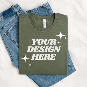 Könnte beinhalten: Ein gefaltetes grünes T-Shirt mit dem Text "YOUR DESIGN HERE" in weißen Buchstaben und weißen Sternen auf weißem Hintergrund. Das T-Shirt liegt auf einer blauen Jeans.