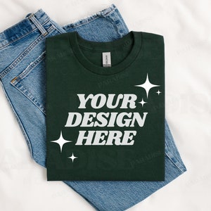 May include: A dark green t-shirt with the text "YOUR DESIGN HERE" in white lettering and small white stars. The t-shirt is folded and placed on top of a pair of blue jeans.