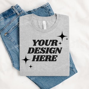 May include: A gray t-shirt with the text "YOUR DESIGN HERE" in black, with small black stars on the sides. The t-shirt is folded and placed on top of a pair of blue jeans.