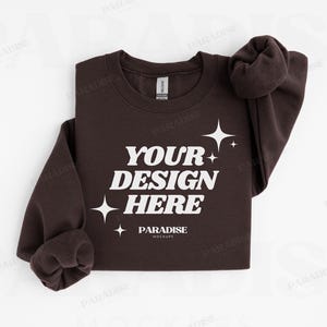 May include: A dark brown sweatshirt with the text "YOUR DESIGN HERE" in white, along with star graphics. The sleeves are cuffed, and the mock-up is on a white background. The brand name "PARADISE MOCKUPS" is also visible.