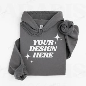 Può includere: Una felpa con cappuccio grigia con una stampa bianca che dice "YOUR DESIGN HERE" e piccole stelle bianche. La felpa è piegata e il cappuccio è tirato su.