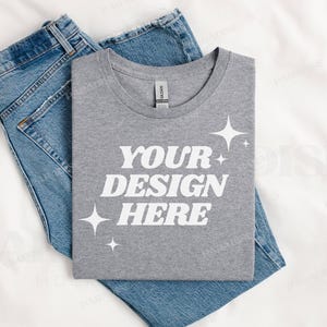 May include: A heather grey t-shirt with the text "YOUR DESIGN HERE" in white, surrounded by small white stars. The t-shirt is folded and placed on top of a pair of blue jeans.