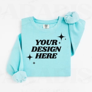 Könnte beinhalten: Ein hellblauer Pullover mit dem Text "YOUR DESIGN HERE" in schwarzer Schrift. Der Pullover hat einen Rundhalsausschnitt und lange Ärmel. Um den Text herum befinden sich kleine Sternchen.