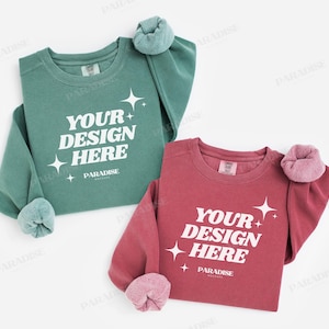 May include: Two crewneck sweatshirts, one teal and one maroon, with the text "YOUR DESIGN HERE" in white. Each sweatshirt has a rolled cuff and the word "PARADISE" below the text. The sweatshirts are on a white background.