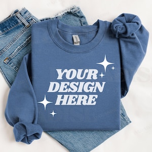 Può includere: Una felpa blu con il testo "YOUR DESIGN HERE" in lettere bianche e piccole stelle bianche. La felpa è piegata e appoggiata su un paio di jeans blu.