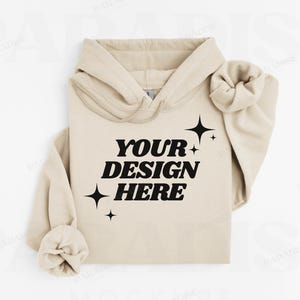 May include: A beige hoodie with the text "YOUR DESIGN HERE" in bold black letters, accented by black starburst graphics. The hoodie features a drawstring hood and cuffed sleeves, perfect for custom designs.
