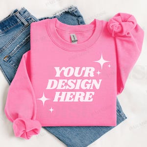 以下が含まれることがあります： 白い文字で「YOUR DESIGN HERE」と書かれたピンク色のスウェットシャツ。ジーンズの上に置かれています。
