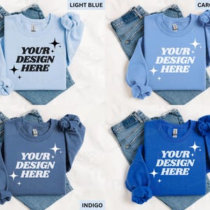 May include: Four crewneck sweatshirts in light blue, Carolina blue, indigo, and royal blue, each with the text "YOUR DESIGN HERE" in white. The sweatshirts are displayed on top of folded denim jeans.