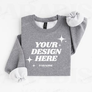 May include: A gray sweatshirt with white text that reads "YOUR DESIGN HERE" and the word "PARADISE" below it. The sleeves are cuffed, revealing a white interior. The sweatshirt is folded neatly.