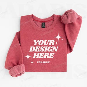 May include: A red crewneck sweatshirt with the text "YOUR DESIGN HERE" in white, along with star graphics. The sleeves are cuffed, and the garment is displayed on a white background. The brand name "PARADISE MOCKUPS" is at the bottom.