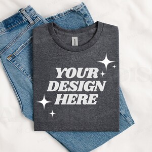 May include: A dark gray t-shirt with the text "YOUR DESIGN HERE" in white lettering and small white stars. The t-shirt is folded and placed on top of a pair of blue jeans.