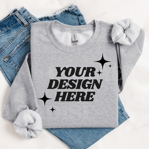 Peut inclure: Un sweat-shirt gris avec le texte "YOUR DESIGN HERE" en lettres noires et de petites étoiles noires. Le sweat-shirt est plié et posé sur une surface blanche avec un jean bleu.