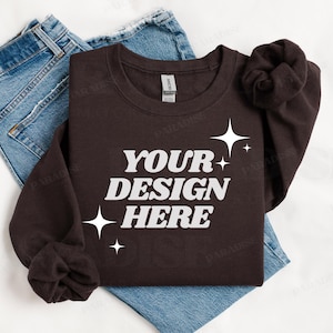 May include: A brown long-sleeve sweatshirt with the text "YOUR DESIGN HERE" in white lettering and a few white stars. The sweatshirt is folded and laid on top of a pair of blue jeans.