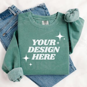 May include: A teal green sweatshirt with the text "YOUR DESIGN HERE" in white. The sweatshirt is folded and laid on a white surface with a pair of blue jeans.