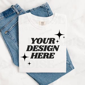 May include: White t-shirt with the text "YOUR DESIGN HERE" in black, surrounded by small black stars. The t-shirt is laid on a white surface with a pair of blue jeans folded on top.
