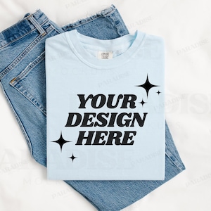 May include: A light blue t-shirt with the text "YOUR DESIGN HERE" in black lettering and small black stars. The t-shirt is laying on a white surface with a pair of blue jeans.