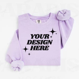 Könnte beinhalten: Ein lavendelfarbener Pullover mit langen Ärmeln und einem Rundhalsausschnitt. Der Pullover hat den Text "YOUR DESIGN HERE" in Schwarz auf der Vorderseite gedruckt, zusammen mit ein paar kleinen Sternen.