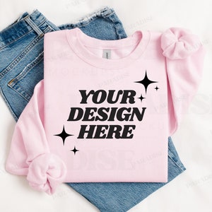 May include: A pink sweatshirt with the text "YOUR DESIGN HERE" in black lettering and small black stars. The sweatshirt is folded and laid on a white surface with a pair of blue jeans.