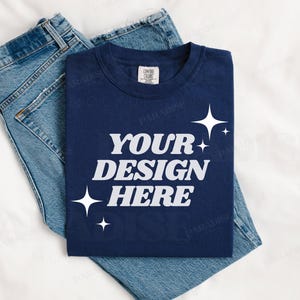 Pode incluir: Uma camiseta azul marinho com o texto "YOUR DESIGN HERE" em branco com estrelas brancas sobre um fundo branco. A camiseta está dobrada e colocada em cima de um par de calças de ganga azuis.
