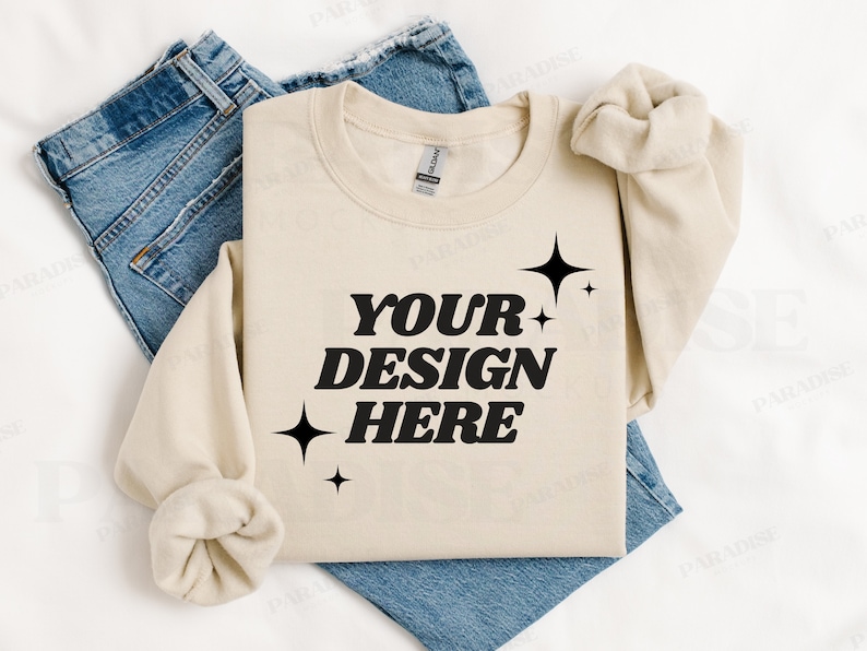 May include: A beige sweatshirt with the text "YOUR DESIGN HERE" in black lettering and small black stars. The sweatshirt is folded and laid on top of a pair of blue jeans.