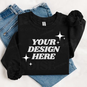 May include: Black sweatshirt with the text "YOUR DESIGN HERE" in white, surrounded by small white stars. The sweatshirt is folded and laid on a white surface with a pair of blue jeans.