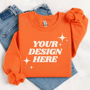 May include: Orange sweatshirt with the text "YOUR DESIGN HERE" in white lettering and small white stars. The sweatshirt is folded and laid on a white surface with a pair of blue jeans.