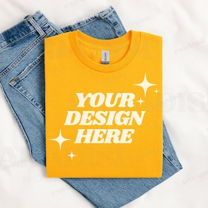 May include: A yellow t-shirt with the text "YOUR DESIGN HERE" in white letters. The shirt is folded and placed on top of a pair of blue jeans.