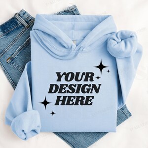 Op de afbeelding: Een lichtblauwe hoodie met een zwarte afbeelding waarop "YOUR DESIGN HERE" staat met sterren rond de tekst.