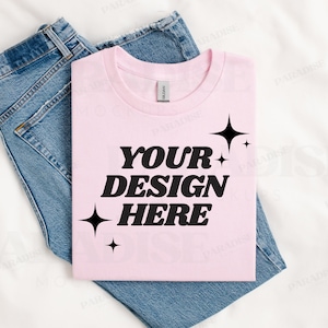 May include: A pink t-shirt with the text "YOUR DESIGN HERE" in black, surrounded by small black stars. The t-shirt is folded and placed on top of a pair of blue jeans.
