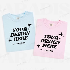 May include: Two folded t-shirts, one light blue and one pink, with the text "YOUR DESIGN HERE" in bold black font. Each shirt has a star graphic and the words "PARADISE MOCKUPS" below the main text.