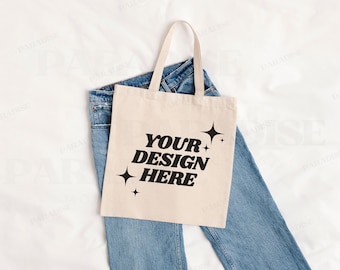 May include: A beige canvas tote bag with black text that reads "YOUR DESIGN HERE" and small star designs. The bag is lying on top of a pair of blue jeans.