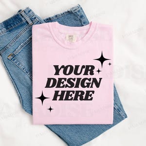 Pode incluir: Uma t-shirt rosa claro com o texto "YOUR DESIGN HERE" em letras pretas em negrito, adornada com estrelas. A t-shirt dobrada está colocada em cima de um par de calças de ganga azuis.
