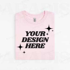 May include: A pink t-shirt with the text "YOUR DESIGN HERE" in black. The text is in a retro font style. There are small black stars around the text.