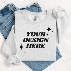 May include: A gray sweatshirt with the text "YOUR DESIGN HERE" in black, surrounded by small stars. The sweatshirt is folded and laid on top of a pair of blue jeans.