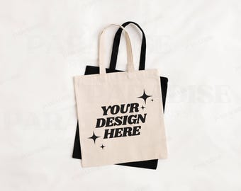 May include: A white tote bag with black handles and the text "YOUR DESIGN HERE" in black. The tote bag is sitting on top of a black tote bag.